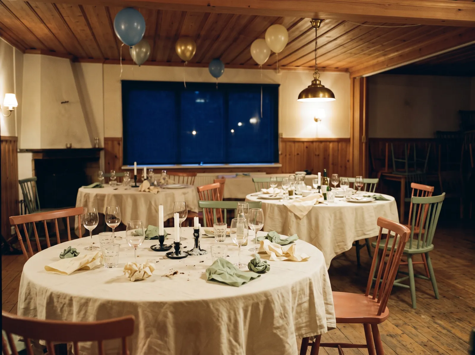 Festlokal en kväll efter festen, med runda bord, utdragna stolar, nedbrunna ljus och ballonger under trätaket.